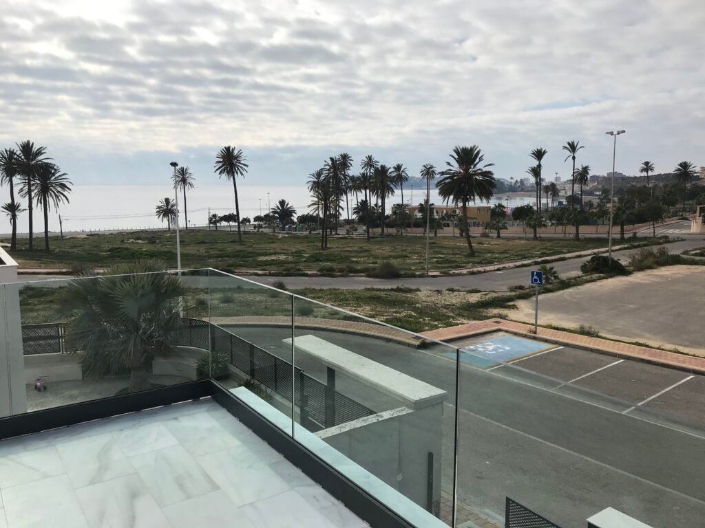 Terraza con barandilla de vidrio y vista al mar con palmeras y paseo marítimo.